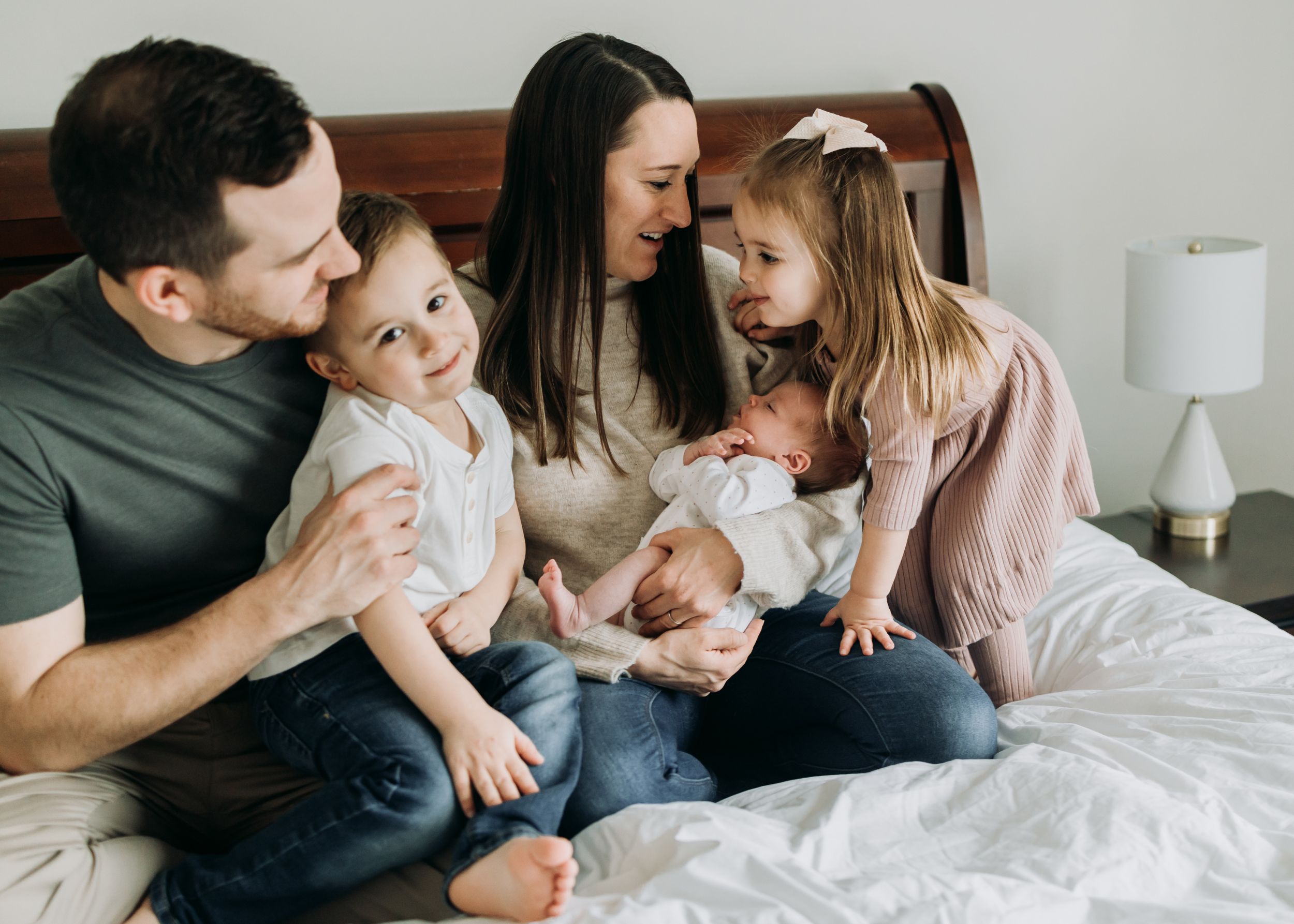 A mom and dad sit on a bed with their toddler son and daughter climbing on them while mom holds their sleeping newborn baby after meeting pittsburgh nannies