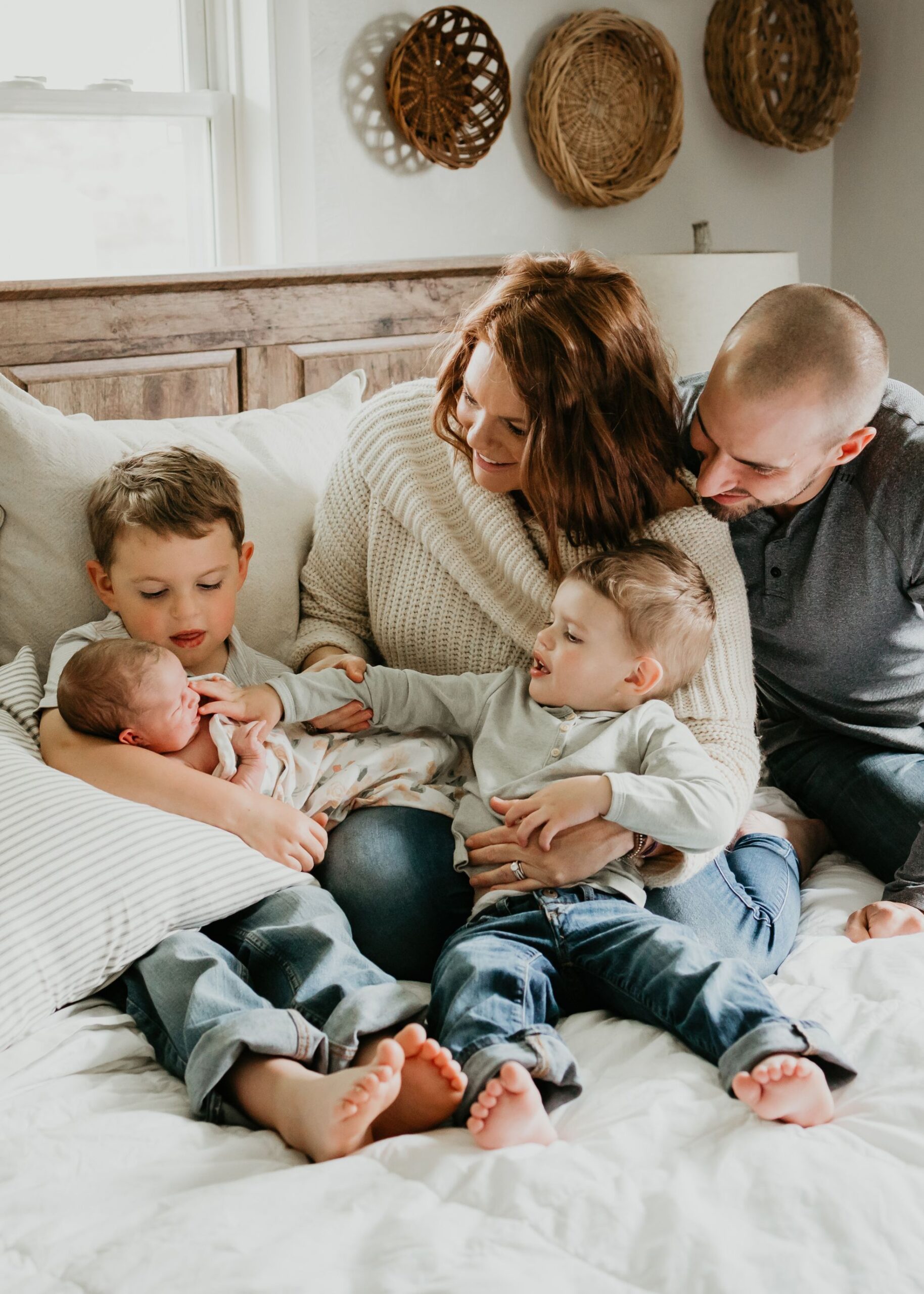 A mom and dad sit on a bed with their two toddler sons while one holds their newborn baby