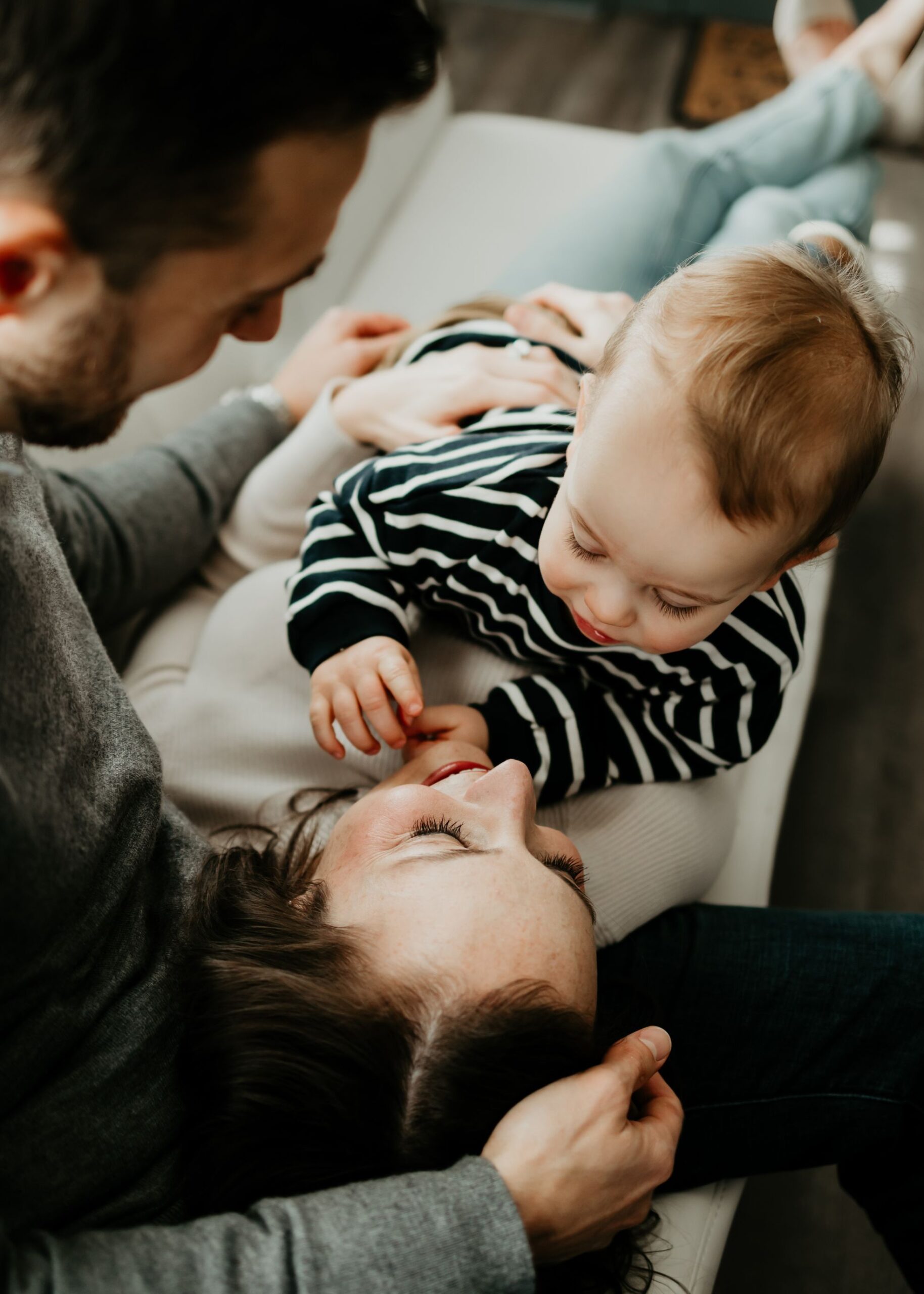 A mother lays on a couch in dad's lap with their toddler playing on her belly after meeting pittsburgh pediatricians