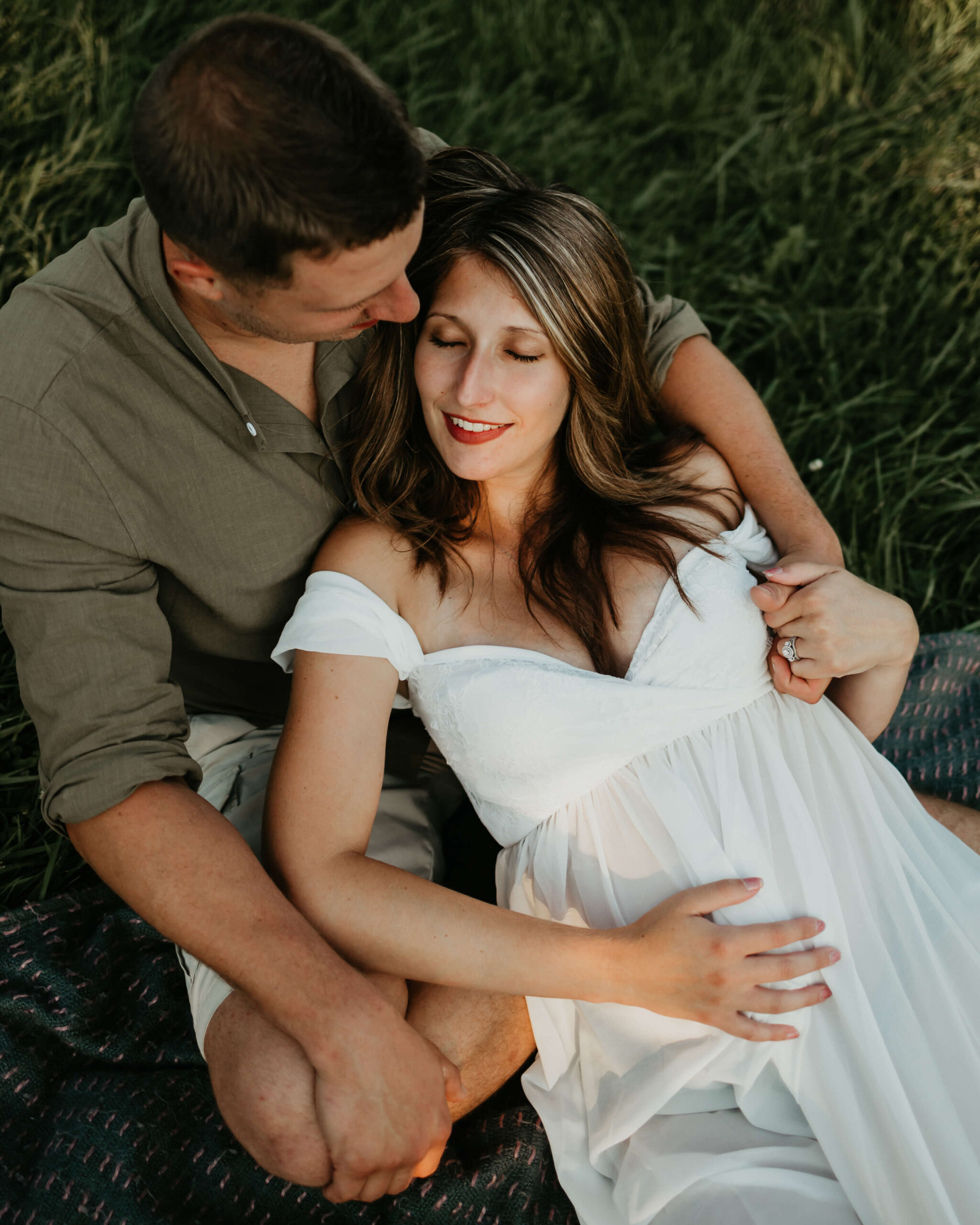 A mom to be lays in the lap of her husband on a picnic blanket in some grass
