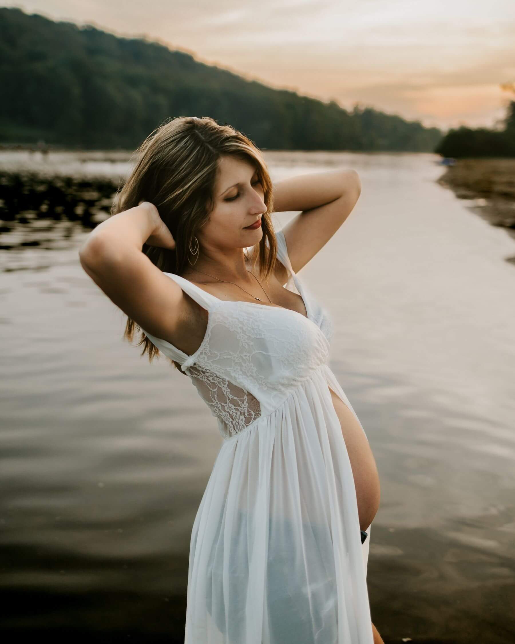 A mother to be in a white open slip with hands in her hair at the edge of the water at sunset after a pittsburgh prenatal massage
