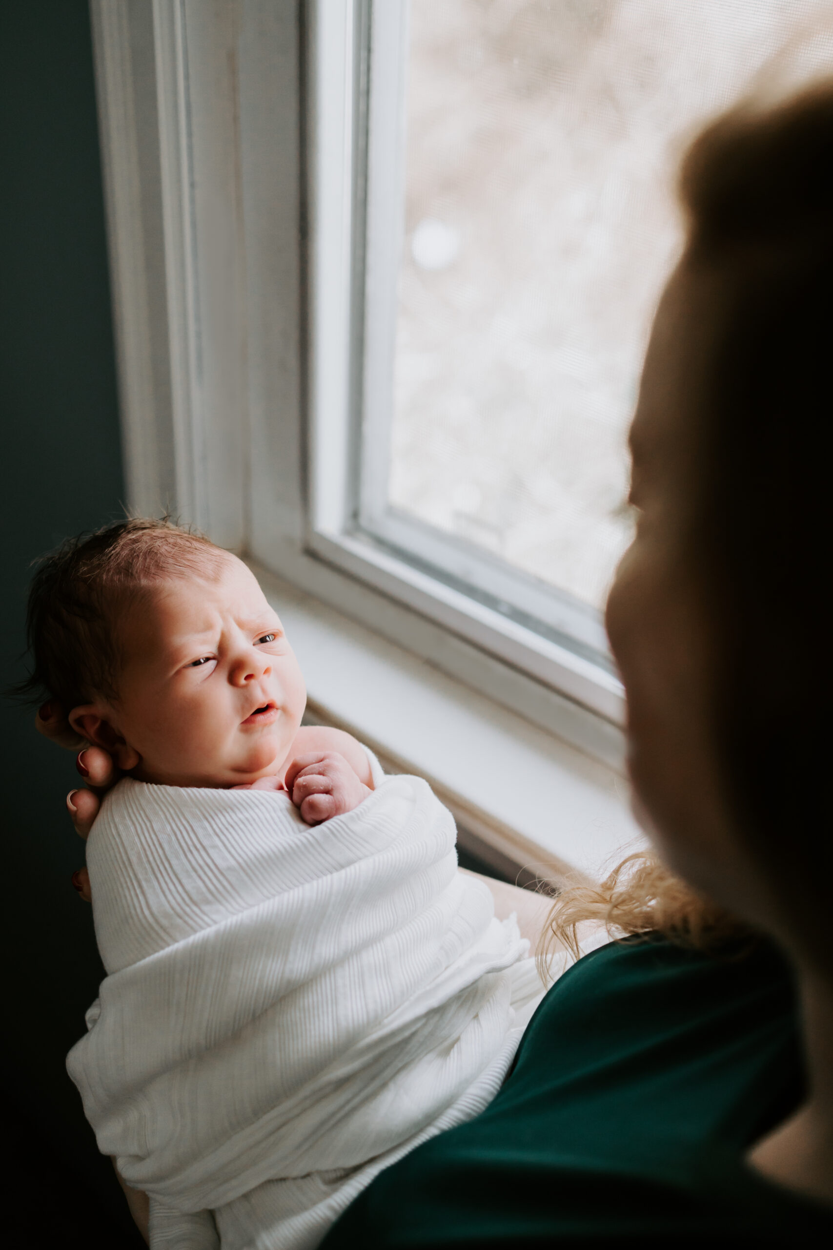 A newborn baby makes a face at mom while she stands in a window after meeting a postpartum doula pittsburgh