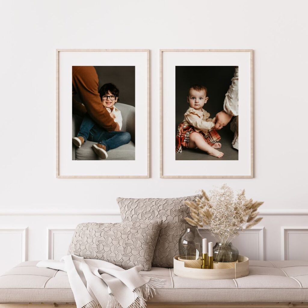 Two framed photos on a wall above a cushioned bench. One shows a child sitting and smiling; the other shows a baby holding hands with a standing person. Dried flowers and candles on the bench.