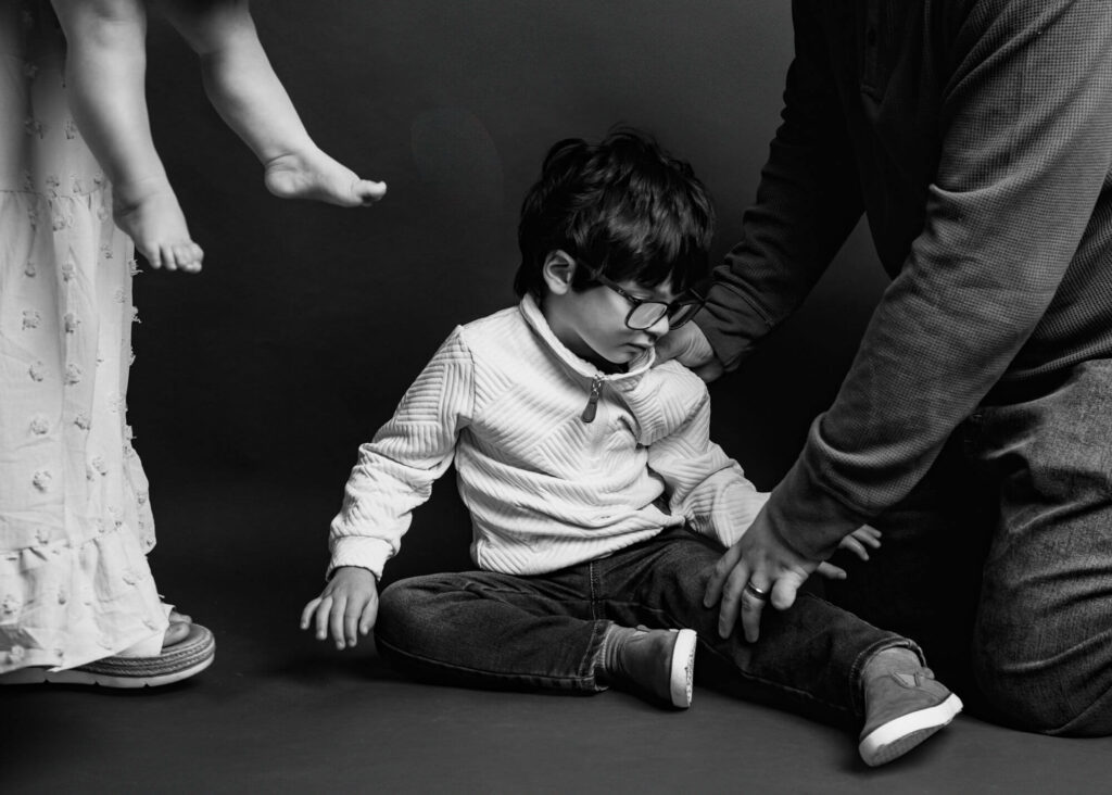 A child wearing glasses sits on the floor. An adult helps support him. Another pair of adult legs is visible nearby during a photoshoot with a sensory friendly photographer in Pittsburgh.