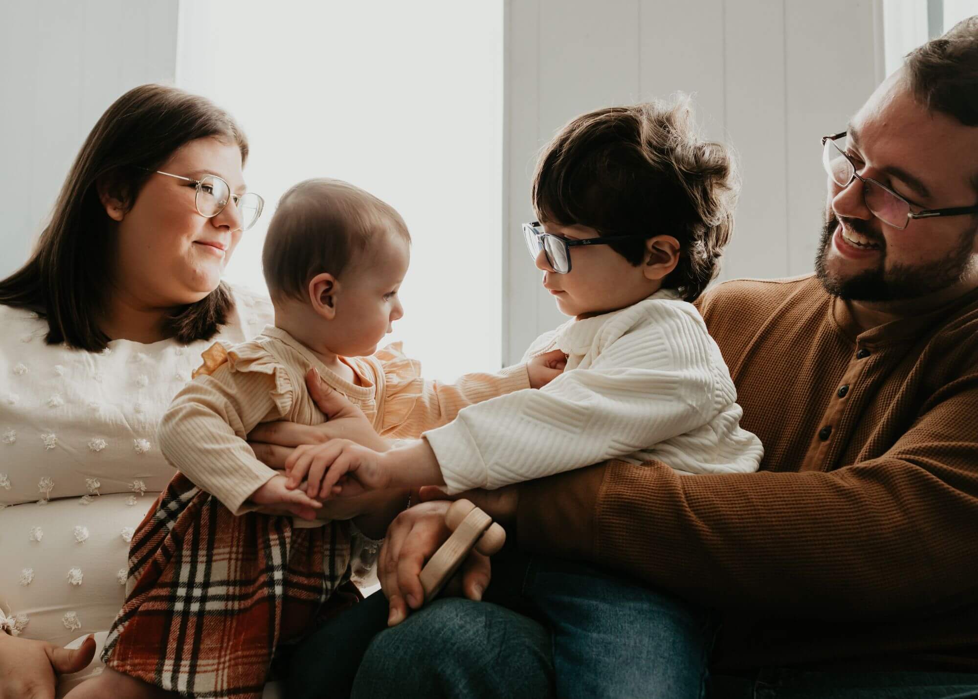 A family of four sitting on a couch. Two adults hold two young children dressed in light-colored clothing. The adults and child on the right wear glasses. They appear to be indoors.