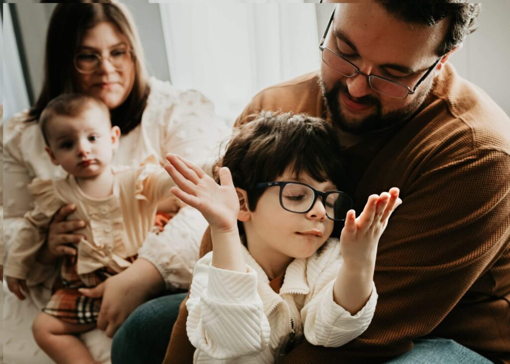 A family of four sitting on a couch. Two adults hold two young children dressed in light-colored clothing. The adults and child on the right wear glasses. They appear to be indoors.