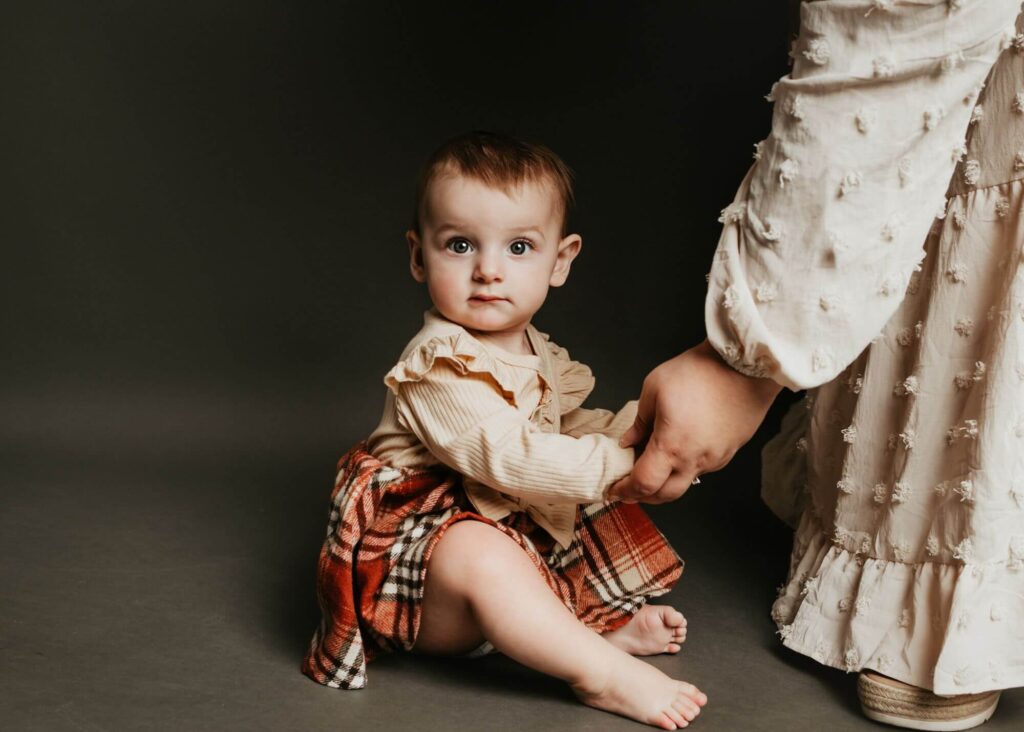 A baby sits on the floor in a Pittsburgh family photography studio holding an adult's hand, wearing a beige top and plaid blanket. The adult is dressed in a textured, light-colored outfit.