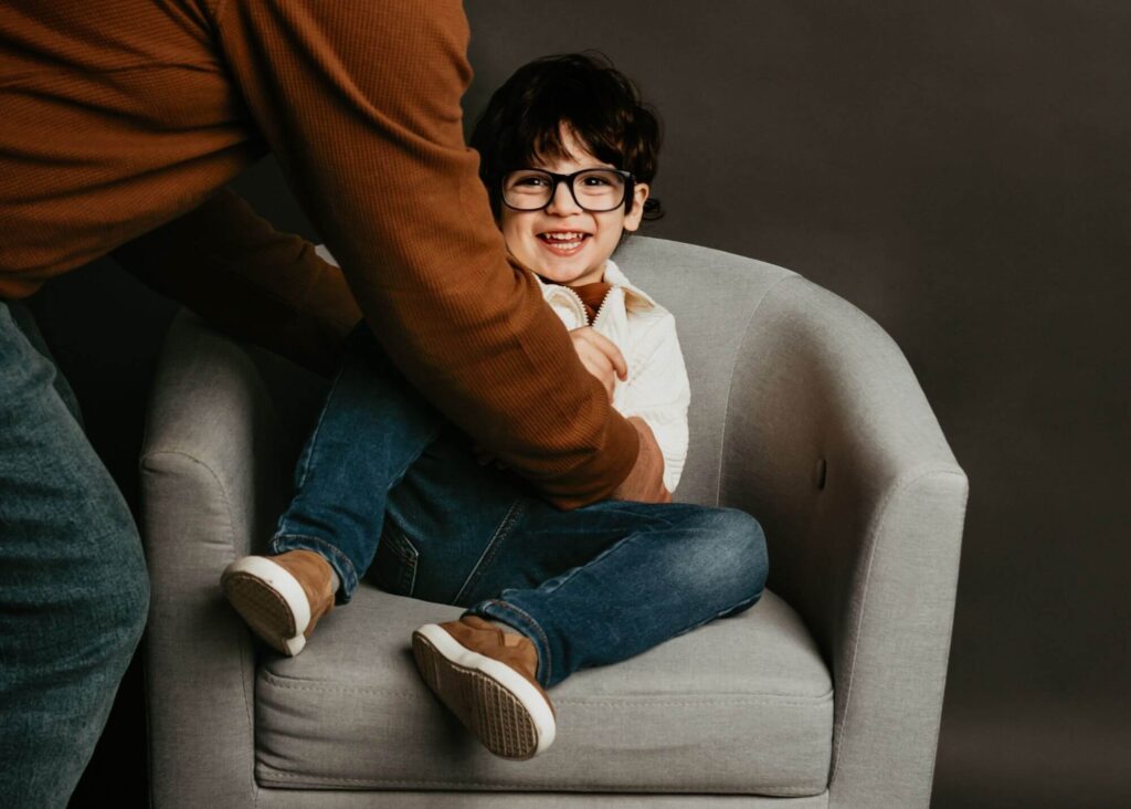 A child with glasses smiles while sitting on a gray armchair, dressed in a white shirt, blue jeans, and brown shoes, holding a toy.