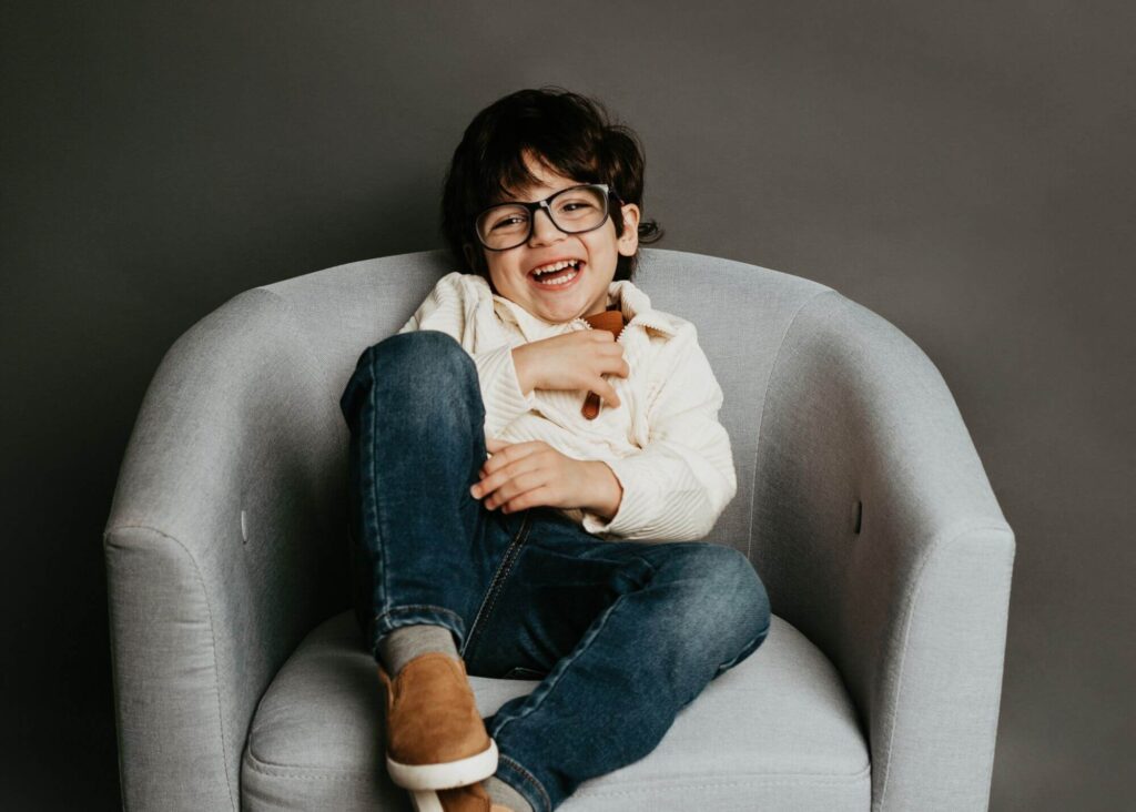 A child with glasses smiles while sitting on a gray armchair, dressed in a white shirt, blue jeans, and brown shoes, holding a toy in a Pittsburgh family photography studio